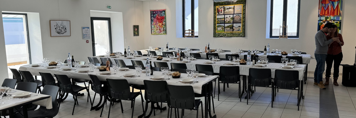 Salle avec tables, chaises, verres, bougies, bouteilles, nappes, tableaux, fenêtres et porte.