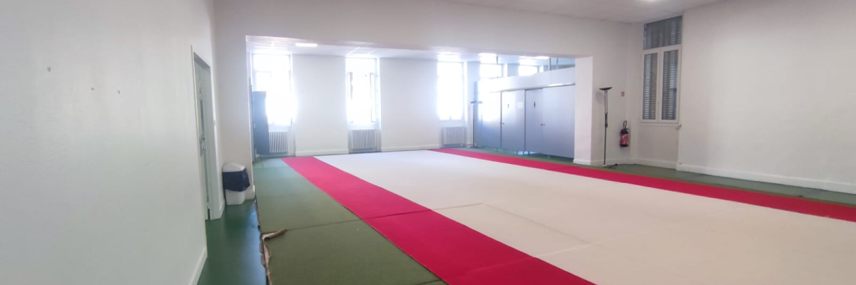 L'image montre une salle de sport vide avec des tapis de sol rouges. Les murs sont blancs, avec des fenêtres et une porte à l'arrière-plan.