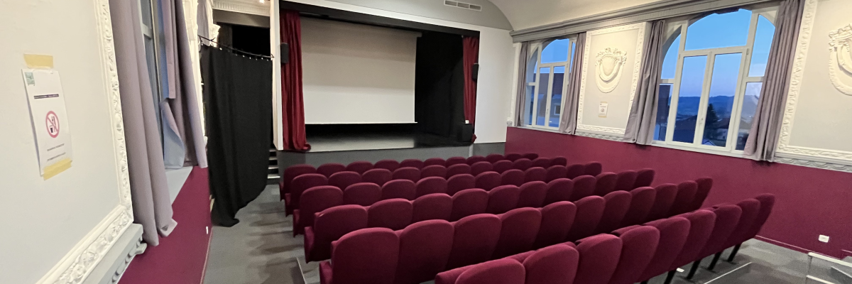 Salle de cinéma avec rangées de sièges rouges, écran, fenêtres et projecteur.