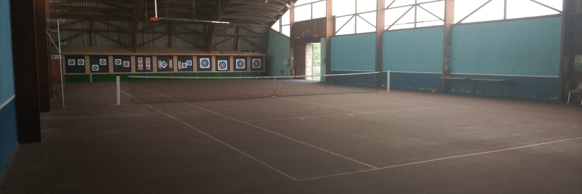 Salle intérieure avec terrain de tennis, filets, cibles de tir à l'arc et éclairage.