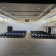 Salle avec rangées de chaises, sol en bois, murs blancs, fenêtres, portes et conduits au plafond.