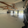 Salle vide avec grandes fenêtres, poutrelles apparentes, spots et ventilateur au plafond.