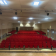 Salle avec rangées de sièges rouges, scène en bois, éclairage au plafond.