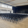 Salle avec rangées de chaises, scène, écran et fenêtres.
