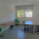 Salle avec tables, chaises vertes, étagère, fenêtre et porte ronde.