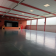 Salle spacieuse avec scène, sol lisse, murs rouges et blancs, grandes fenêtres et éclairage au plafond.