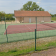 Terrain de tennis avec clôture, filet et banc. Bâtiment adjacent.