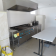 Cuisine avec plaque de cuisson, hotte, réfrigérateur, table et balai.