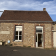 Bâtiment en pierre avec toit en tuiles, porte blanche ouverte, fenêtre à droite, porte de garage blanche à gauche.