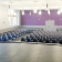 Salle avec rangées de chaises, scène, fenêtres, portes et éclairage suspendu.