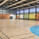 Gymnase avec paniers de basket, filet de handball, gradins, sol en parquet et murs colorés.