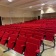 Salle avec rangées de chaises rouges, table à l'avant, écran et porte.