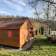 Petite maison en bois avec toiture en tuiles, portes rouges, située dans un jardin avec arbres et autre habitation au loin.