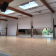 Salle avec sol en bois, murs blancs, poutrelles apparentes, écran, fenêtres et éclairage naturel.