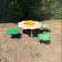 Aire de jeux avec table en forme de fleur, siège jaune et quatre pas en forme de feuilles vertes.