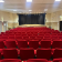Salle avec rangées de sièges rouges, scène, rideau, éclairage et portes.