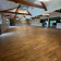 Grande salle avec parquet, poutres apparentes, luminaires suspendus, portes vitrées et fenêtres.