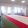 L'image montre une salle de sport vide avec des tapis de sol rouges. Les murs sont blancs, avec des fenêtres et une porte à l'arrière-plan.