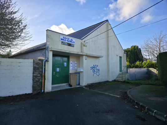 Un petit bâtiment avec un panneau indiquant "Salle de Mez-Kelo". Porte verte, poubelles, graffiti, mur de pierre, allée.