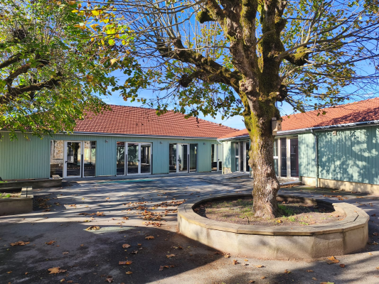 Bâtiment à un étage avec toit rouge, murs verts et portes en verre. Cour avec arbre et cercle de béton autour.