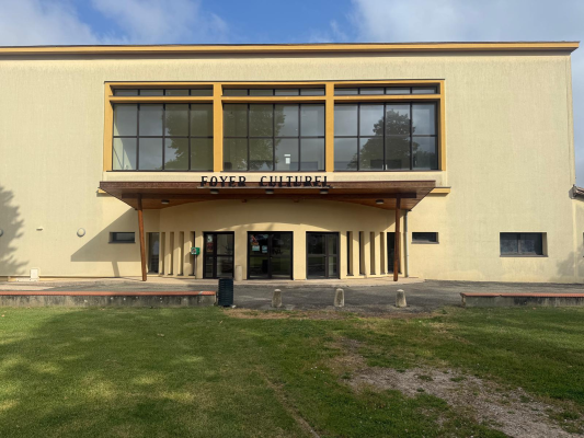 Bâtiment avec entrée, grandes fenêtres, enseigne "Foyer Culturel", pelouse et ciel.