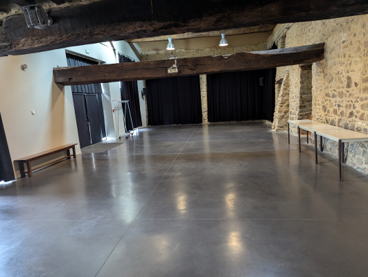 Salle avec tables, bancs, rideaux, sol poli, poutres et pierres apparentes.