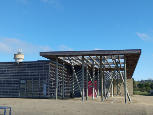 Bâtiment moderne avec structure en bois et façade en verre. Toit en surplomb soutenu par des poteaux métalliques. Ciel bleu en arrière-plan.