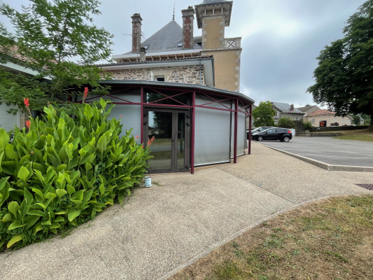 Bâtiment avec extension moderne, allée goudronnée, voiture et buissons.