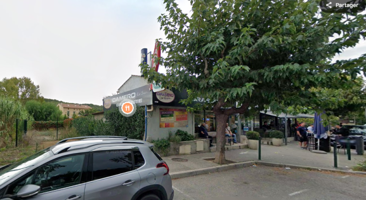 Bâtiment avec enseigne "CAMERO LAVIE" et parking. Voiture devant. Arbre et tables en terrasse.