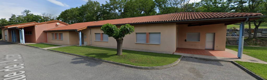 Bâtiment avec toiture en tuiles, murs crème et portes bleues. Arbre et pelouse devant, route et parking à proximité.