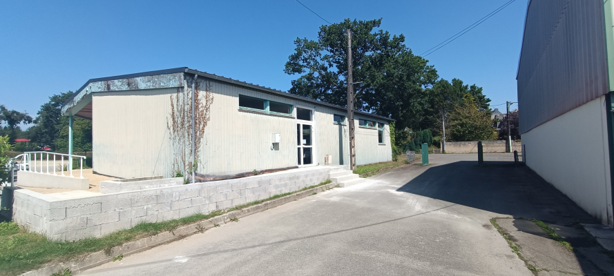 Bâtiment bas avec façade blanche, toit gris, rampes d'accès, portes vitrées, arbres et route asphaltée.