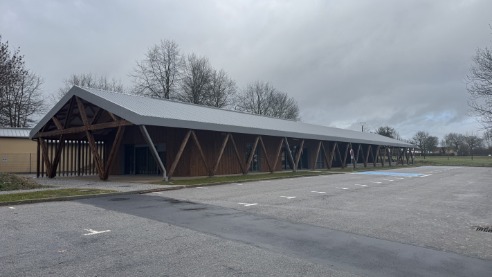Bâtiment long avec toit incliné, structure en bois et grandes fenêtres, stationnement devant.