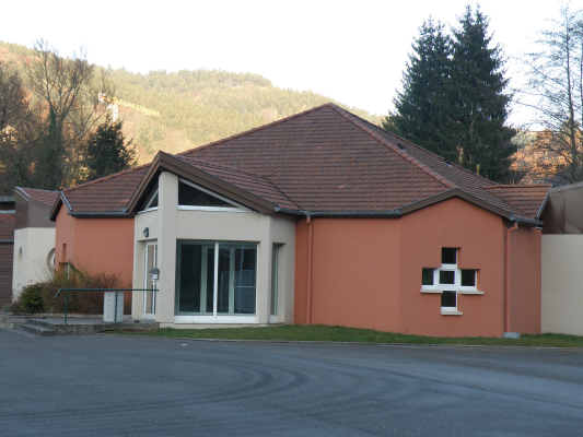 Bâtiment avec toit en pente, murs peints en rose et blanc, fenêtres, porte vitrée, et une croix en brique sur le mur.