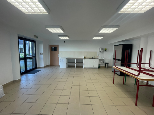 Salle avec tables, chaises, évier, réfrigérateur, et porte. Sol carrelé, lumières au plafond.