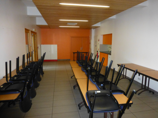 Salle avec tables, chaises, tableau et équipement électrique. Murs orange, sol gris et éclairage intégré au plafond.