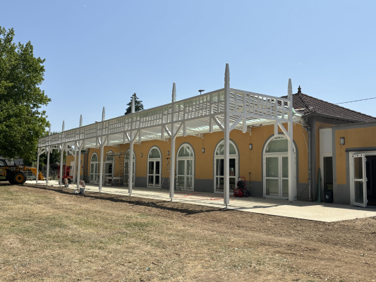 Bâtiment avec structure blanche et murs jaunes, portes vitrées, toit en tuiles, et terrain herbeux.