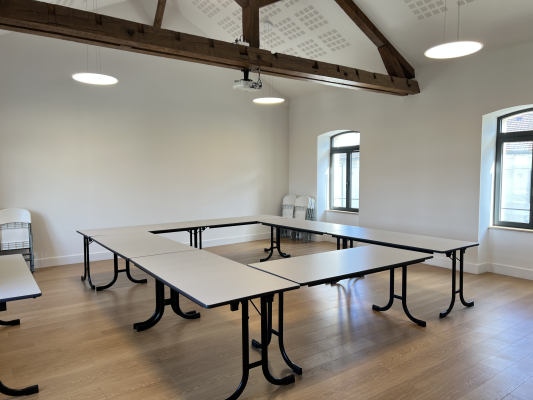 Salle avec tables rectangulaires, chaises, lustres, fenêtres.