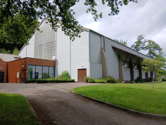 Bâtiment avec façade principale blanche, toiture grise, et extension en brique. Grandes fenêtres, porte centrale, pelouse verte et allée.