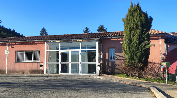 Bâtiment rose avec toiture en tuiles, portes vitrées et fenêtres grillagées. Arbre et panneau de stationnement visible.