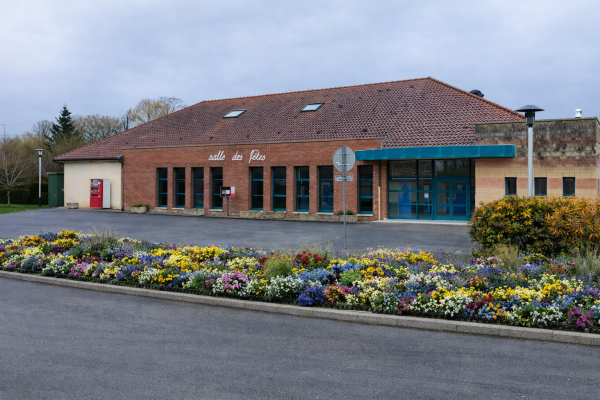 Bâtiment avec toiture en tuiles, murs en brique, grandes fenêtres, entrée avec auvent bleu, panneau de signalisation, parterre de fleurs.