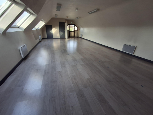 Salle vide avec parquet, fenêtres, radiateurs et éclairages encastrés.