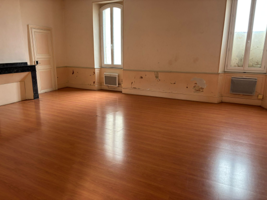 Pièce vide avec parquet, murs endommagés, radiateurs et fenêtres.