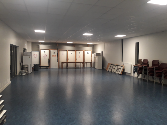 Salle vide avec tables, chaises, portes numérotées, radiateurs et frigo.