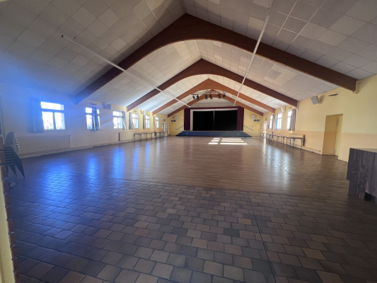 Grande salle avec sol en bois, murs blancs, plafond voûté, fenêtres, rideaux, radiateurs et scène.