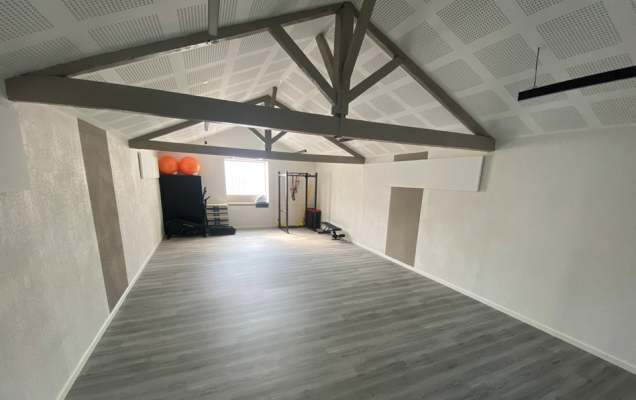 Salle vide avec poutres apparentes, parquet, mur blanc et équipement de sport.