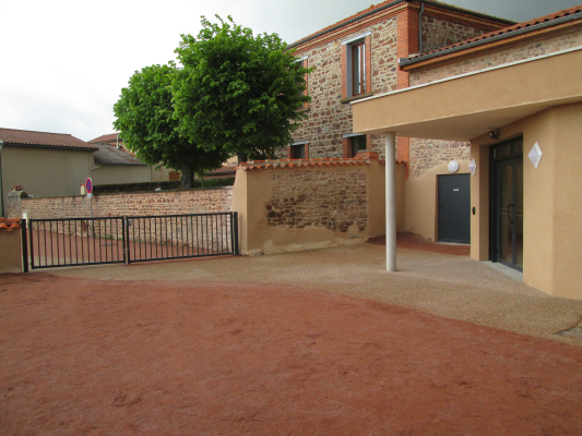 Cour avec sol en terre battue, bâtiment en pierre et briques, portail métallique, arbre.