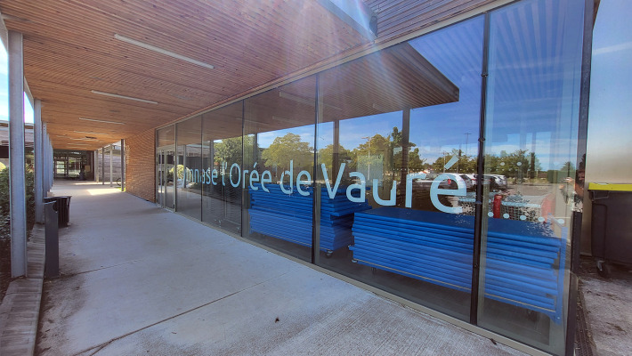 Entrée avec toit, murs vitrés, bancs à l'intérieur, arbres et ciel visibles à travers les vitres.
