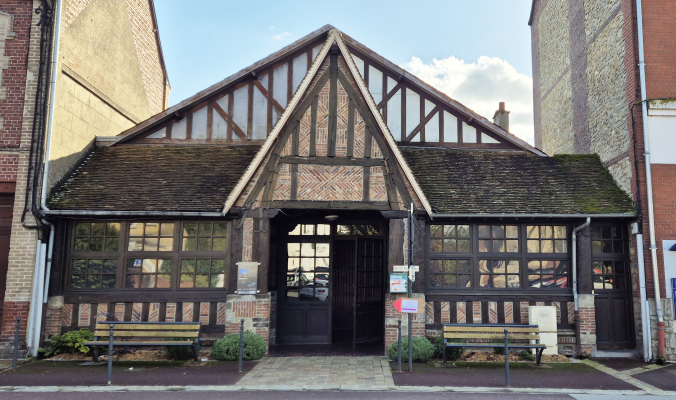 Bâtiment à structure en bois avec toit en tuiles, grandes fenêtres et porte centrale. Deux bancs devant, plantes et panneaux.