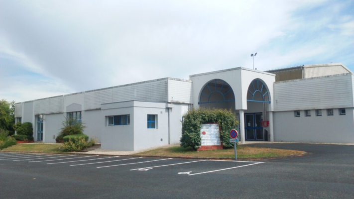 Bâtiment blanc avec entrées, fenêtres, buissons, parking et panneaux de signalisation.