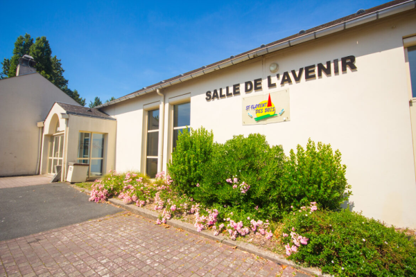 Bâtiment blanc avec enseigne "Salle de l'Avenir". Entrée avec porte vitrée. Buissons et fleurs.